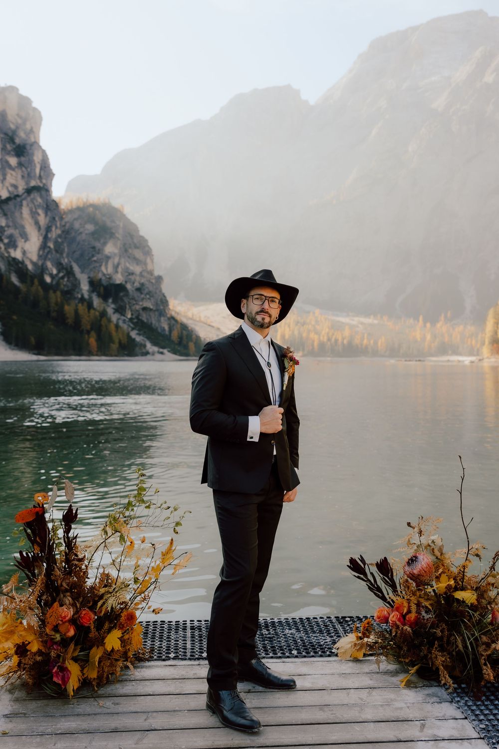 A groom in a black suit and cowboy hat stands on a dock by a mountain lake with autumn floral arrangements.