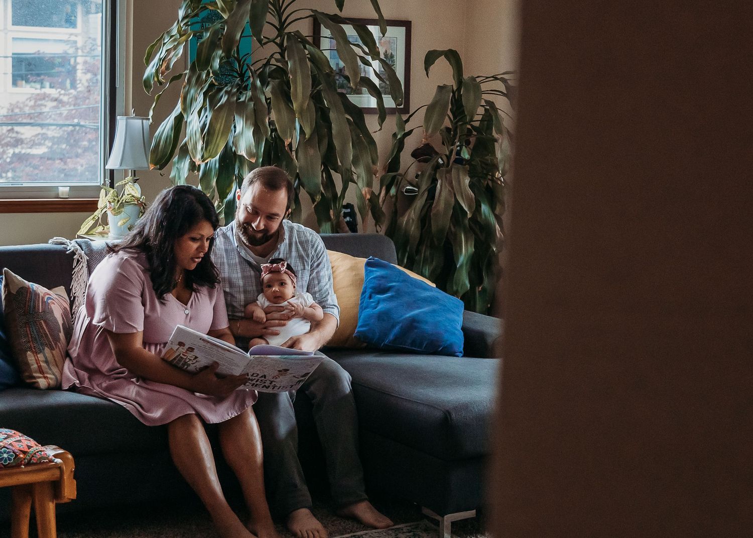 A family sits together on a gray couch in a dimly lit living room with plants in the background.