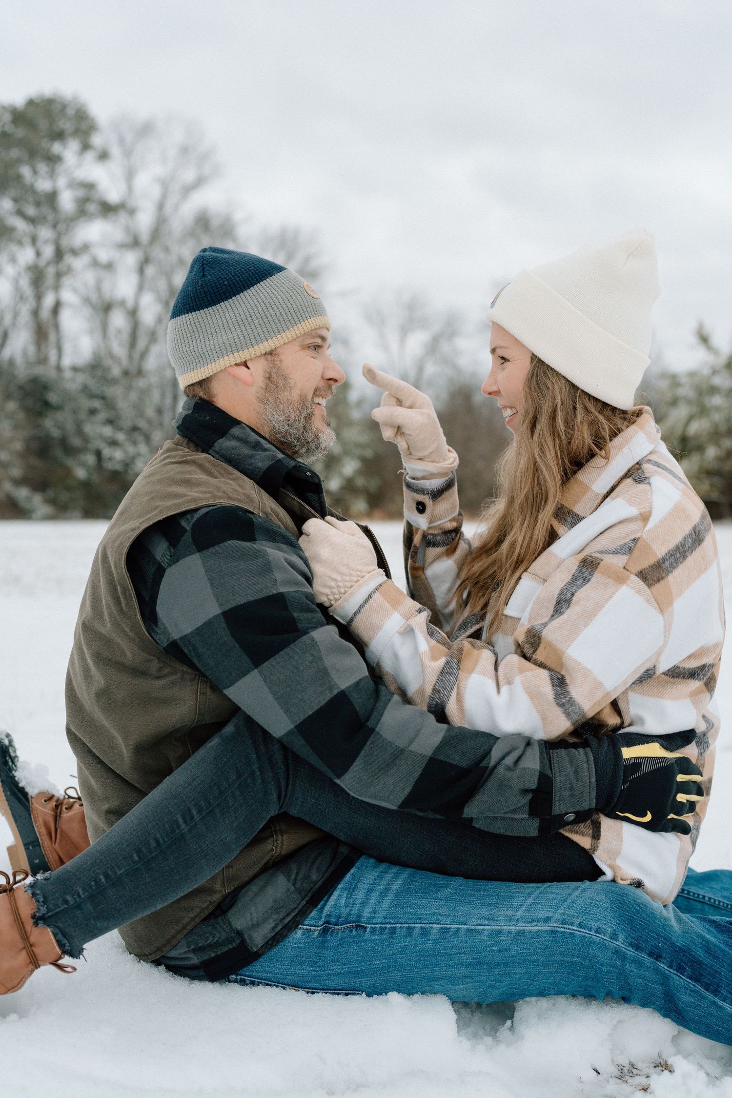 Playful couple lying in snow having fun during winter photoshoot wearing plaid and dark jackets Chattanooga Lifestyle Wedding Photographer
