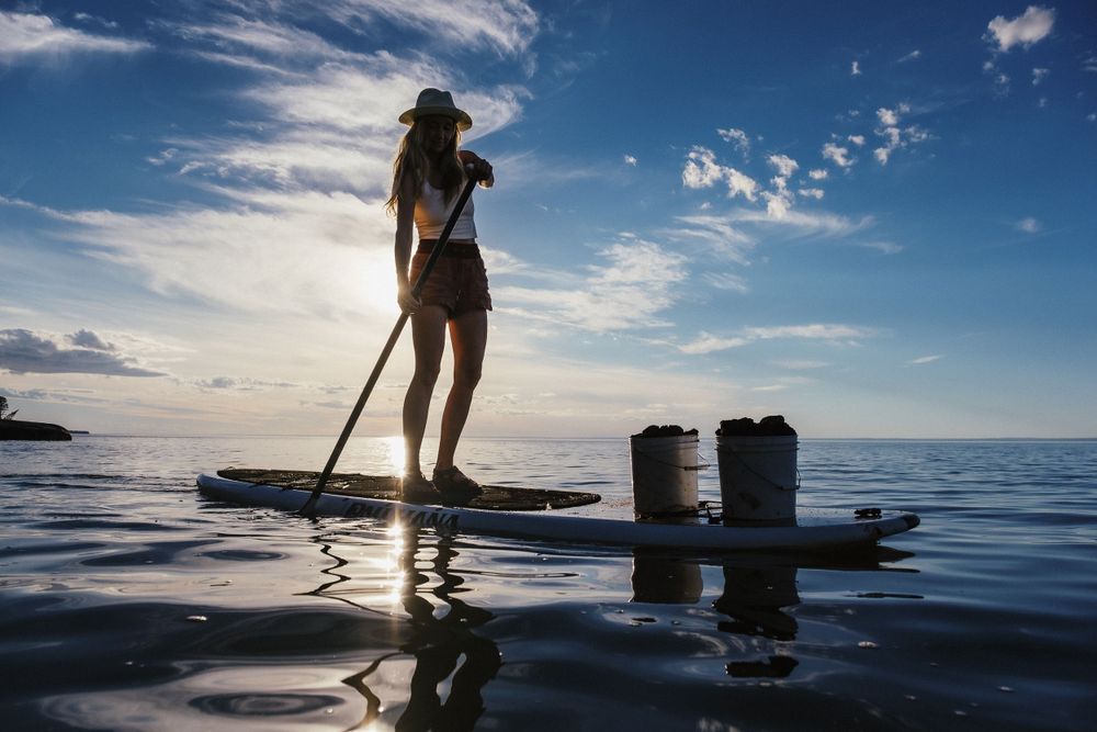Silhouetted paddleboarder stands on calm ocean waters against a bright blue sky with scattered clouds at sunset.