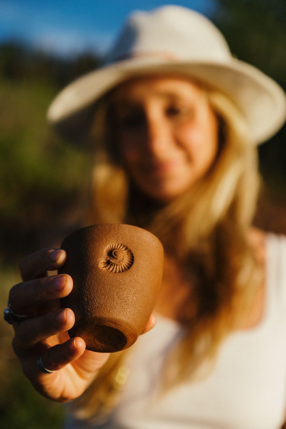 A person wearing a white hat holds up a brown ceramic cup with a spiral design in warm sunset lighting.