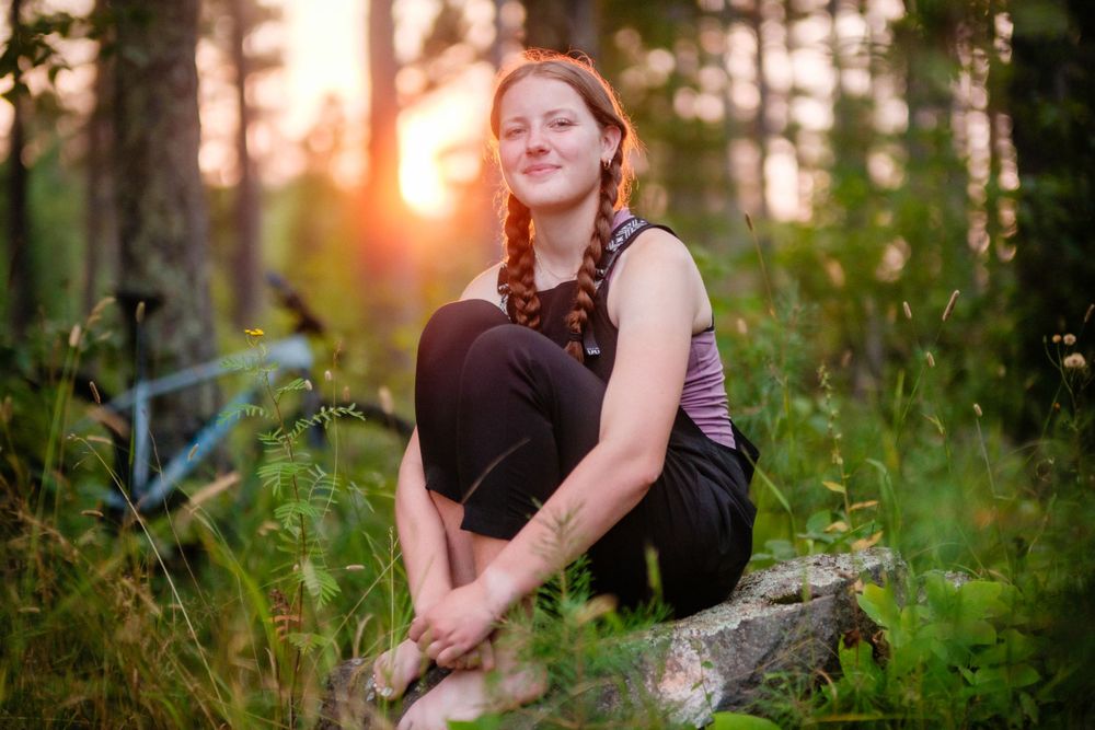 A person in athletic wear sits on a rock in a forest during golden hour sunset, surrounded by greenery.