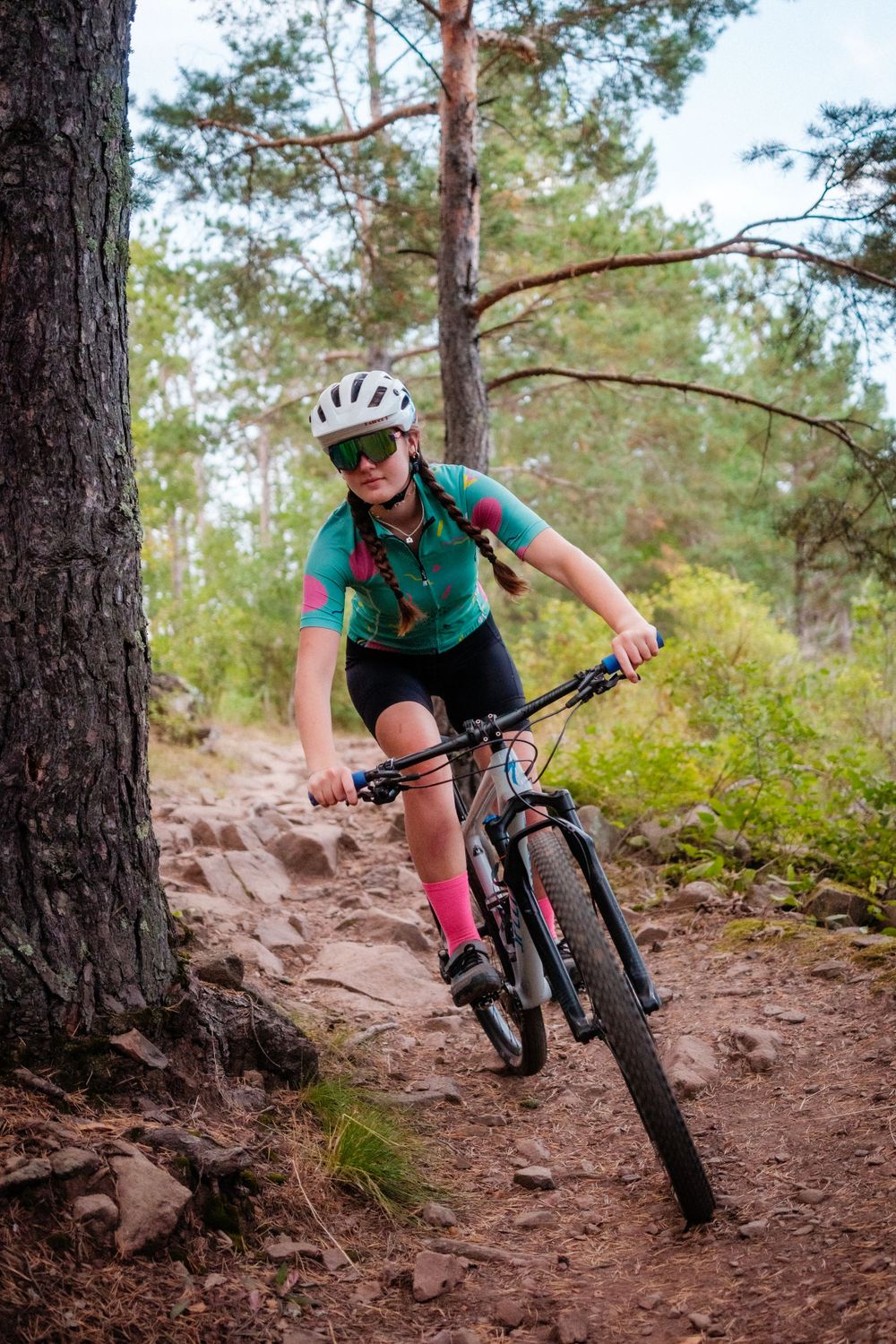 Mountain biker rides down rocky forest trail wearing turquoise jersey and helmet with pink socks and black shorts.