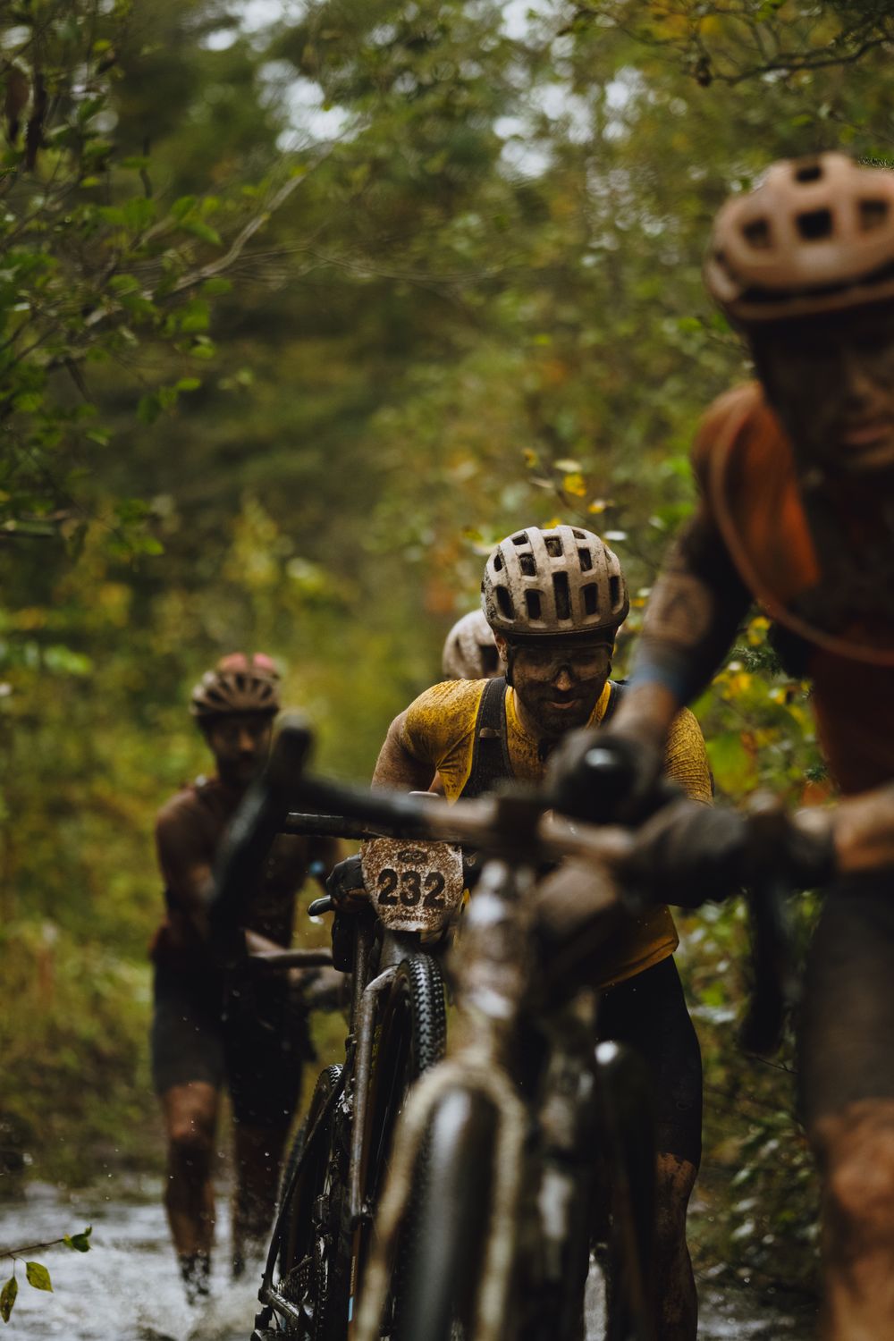 Mountain bikers in helmets riding through wet forest terrain during a rainy trail ride.