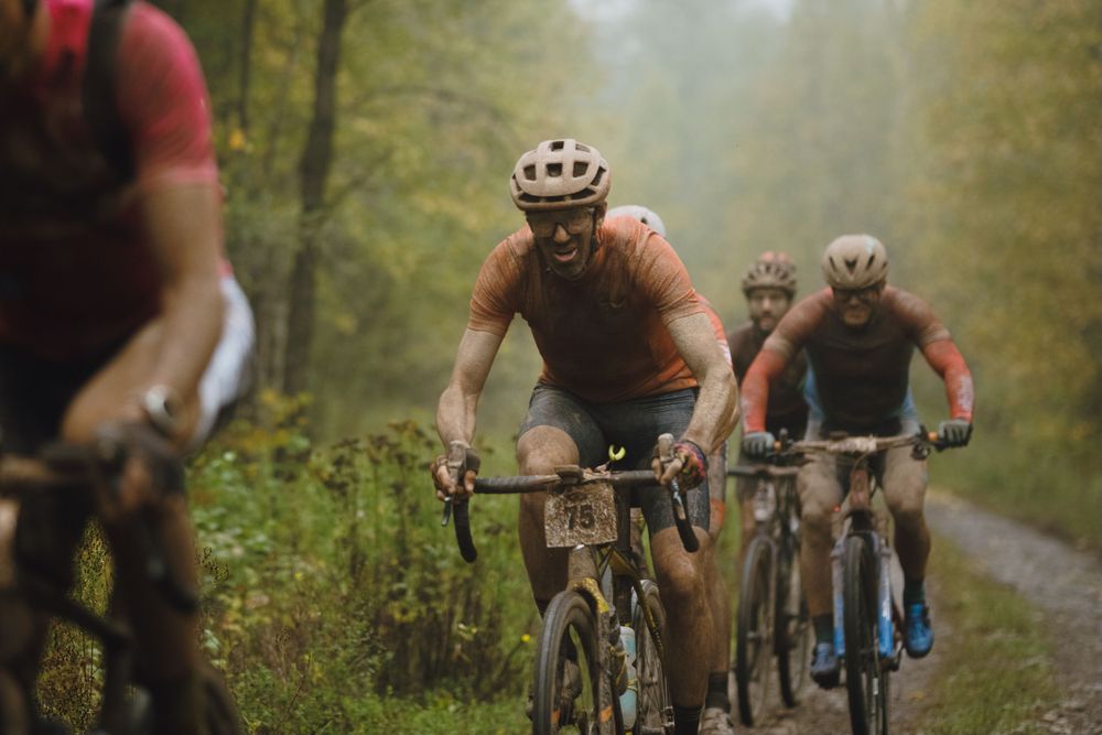 Cyclists riding mountain bikes on a misty forest trail during a group ride along a green wooded path.