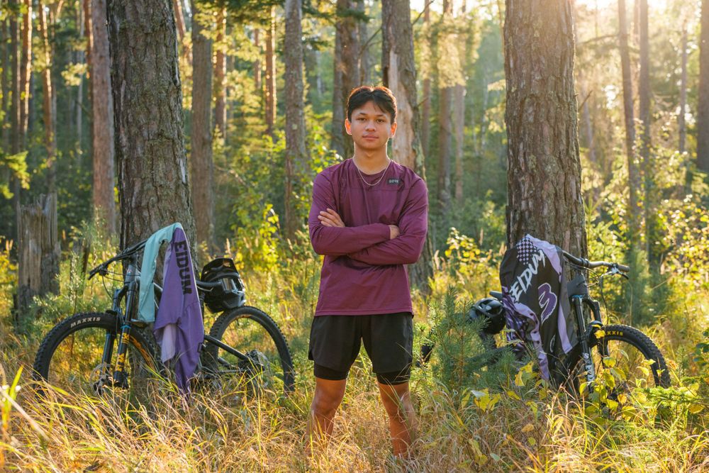 A cyclist stands between two mountain bikes in a sunlit forest with tall pine trees and golden grass.