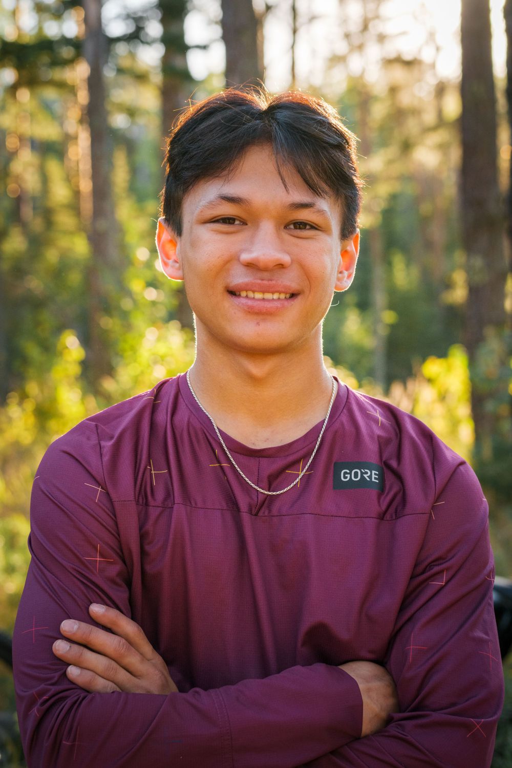 A person wearing a purple Gore-tex jacket poses outdoors with crossed arms in warm sunlit forest.
