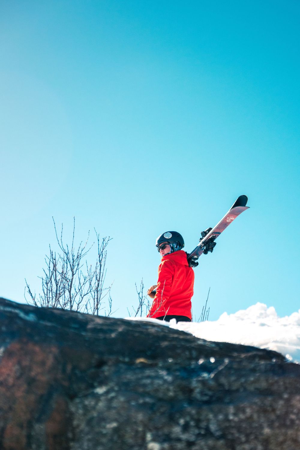 A skier in a red jacket stands atop a snowy cliff edge against a vibrant blue sky.