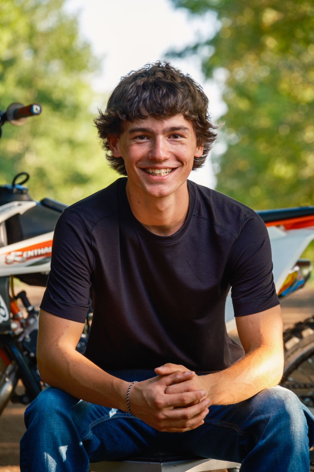 A young person in a black t-shirt sits smiling outdoors with a dirt bike visible in the background.