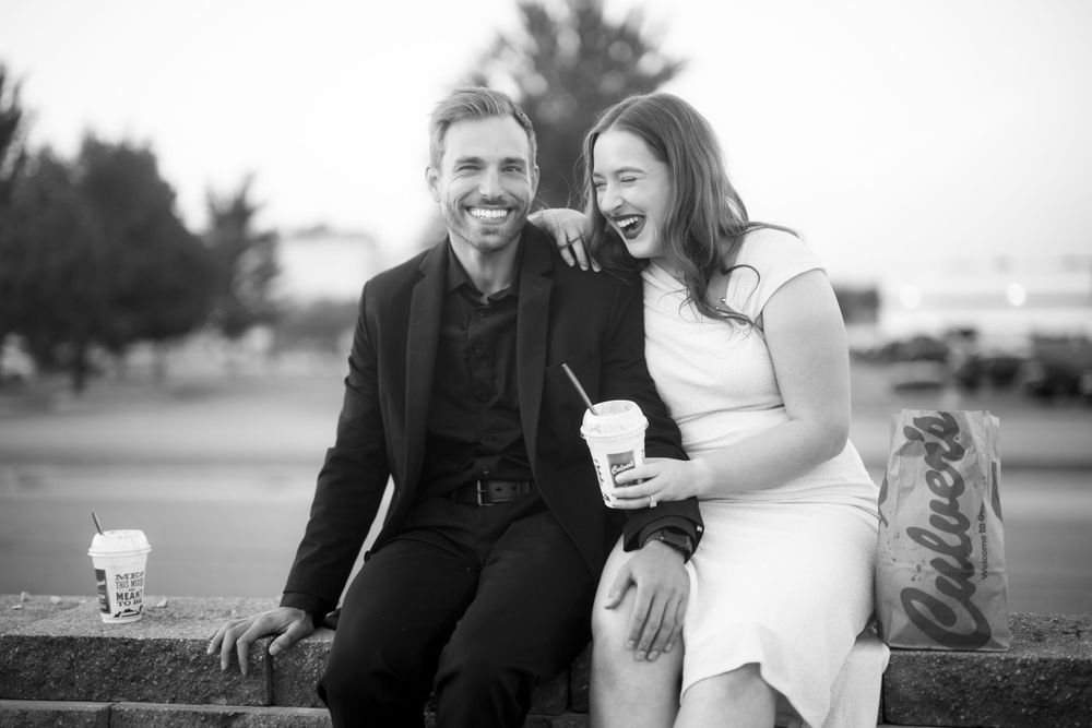A happy couple sits outdoors sharing a laugh while holding coffee cups in black and white photography.