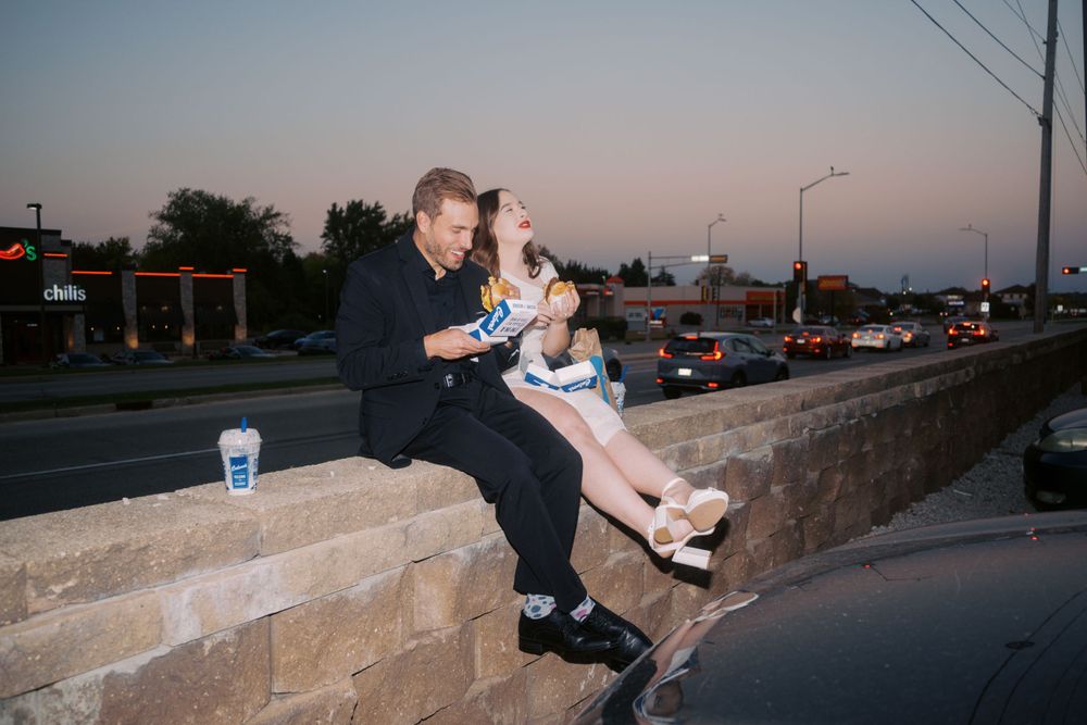 Two people sit together on a stone wall at dusk as traffic passes by on the street below.