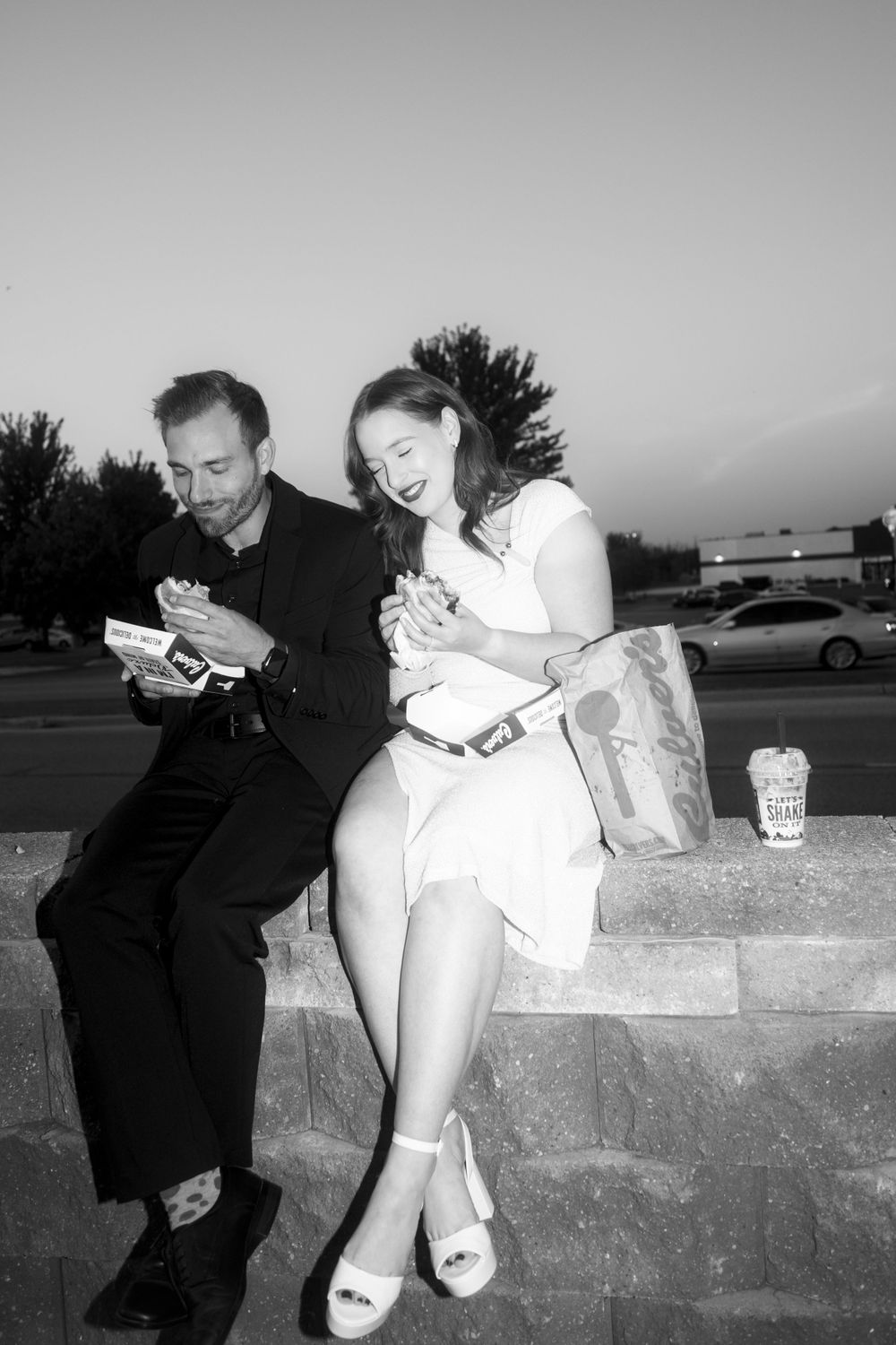 Two people sitting outdoors on stone steps looking at mobile phones while eating takeout food.
