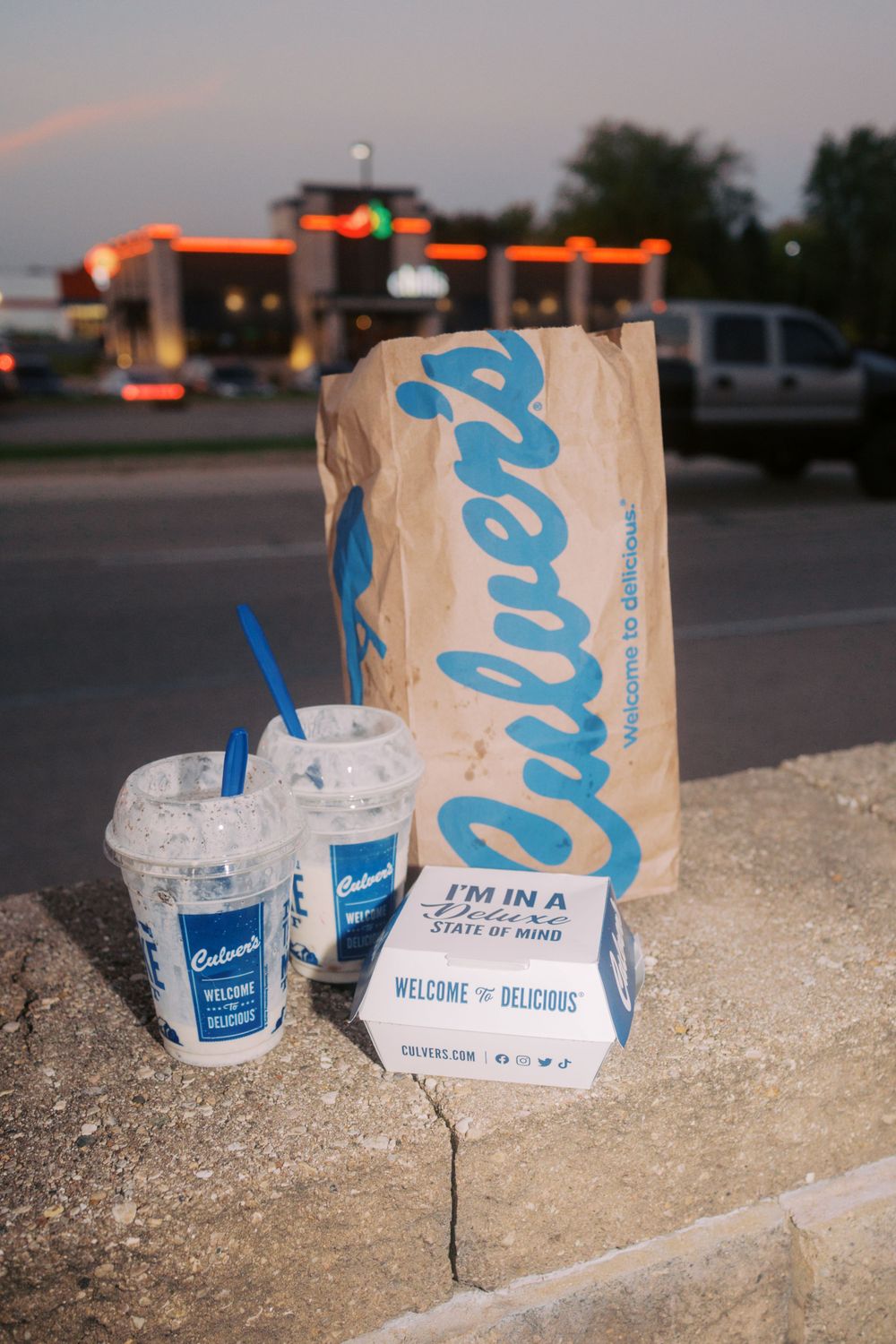 Culver's fast food restaurant takeout bag and drinks sit on concrete ledge at dusk with gas station lights in background.