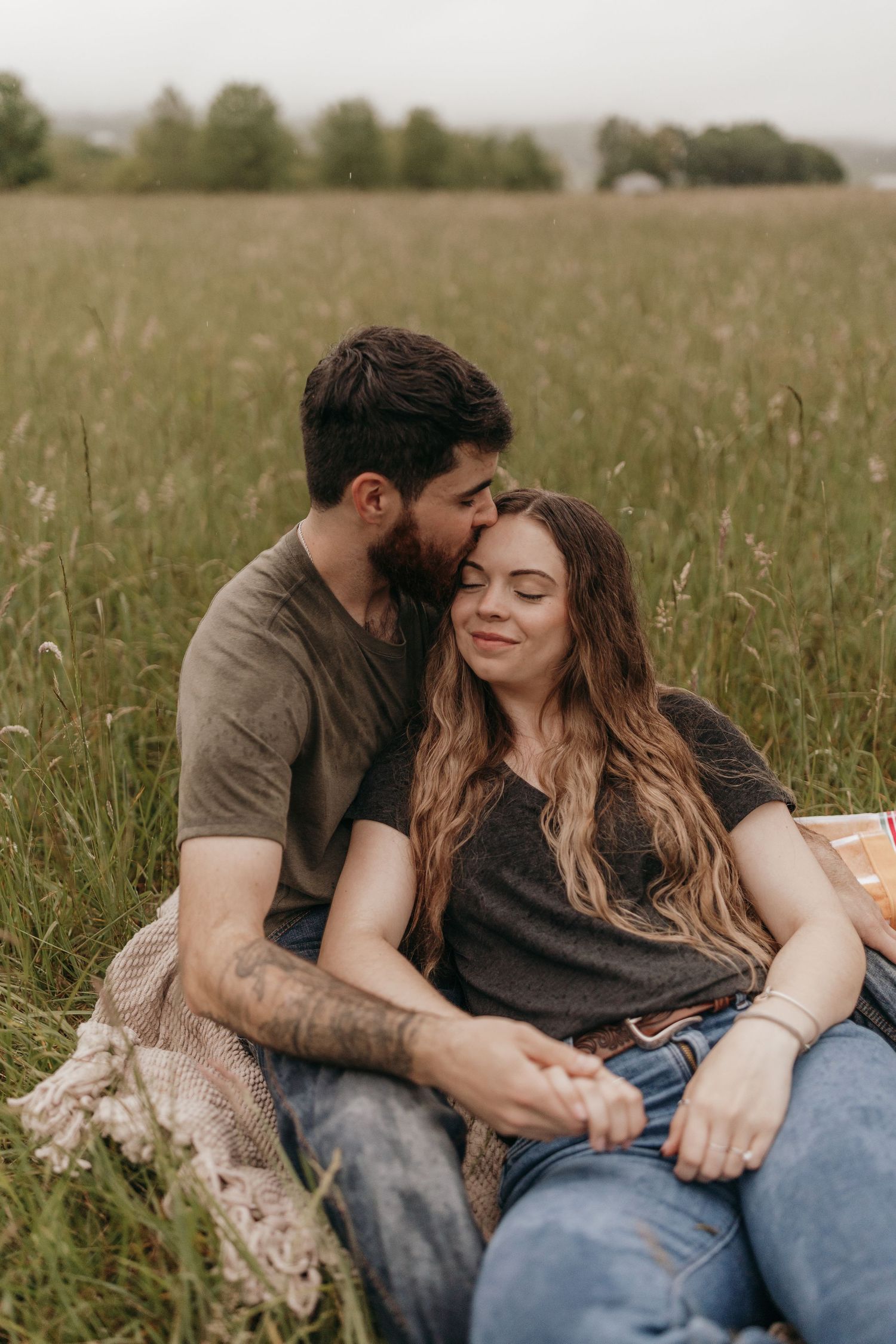 An intimate candid moment between an engaged couple sitting in a rainy, rustic field.