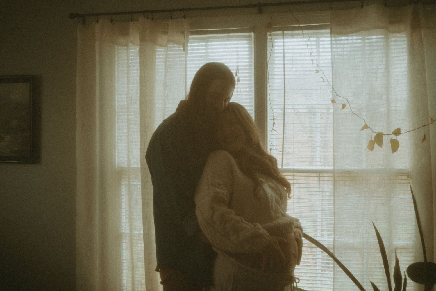 A silhouette of a couple embracing by a sunlit window with sheer curtains.