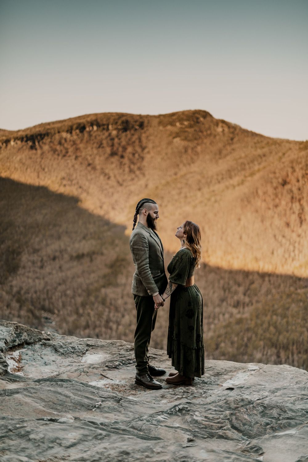 Two figures share an intimate moment on rocky mountain cliff during golden hour.