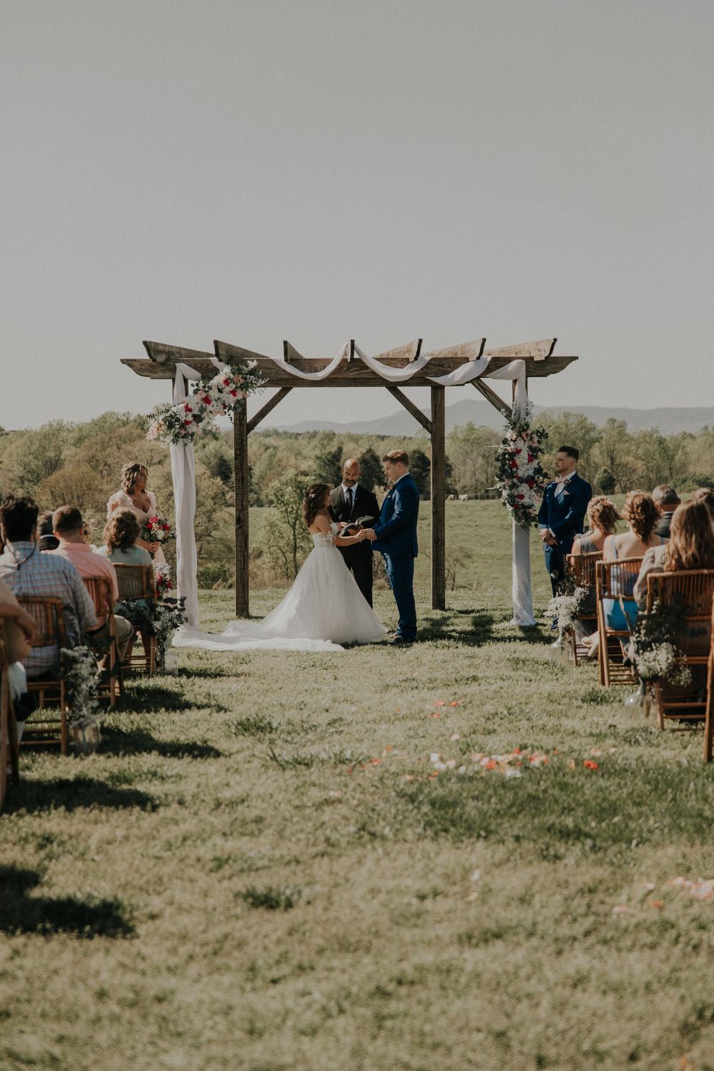 Outdoor wedding ceremony setup with wooden arch and chairs on grassy hillside under clear sky.