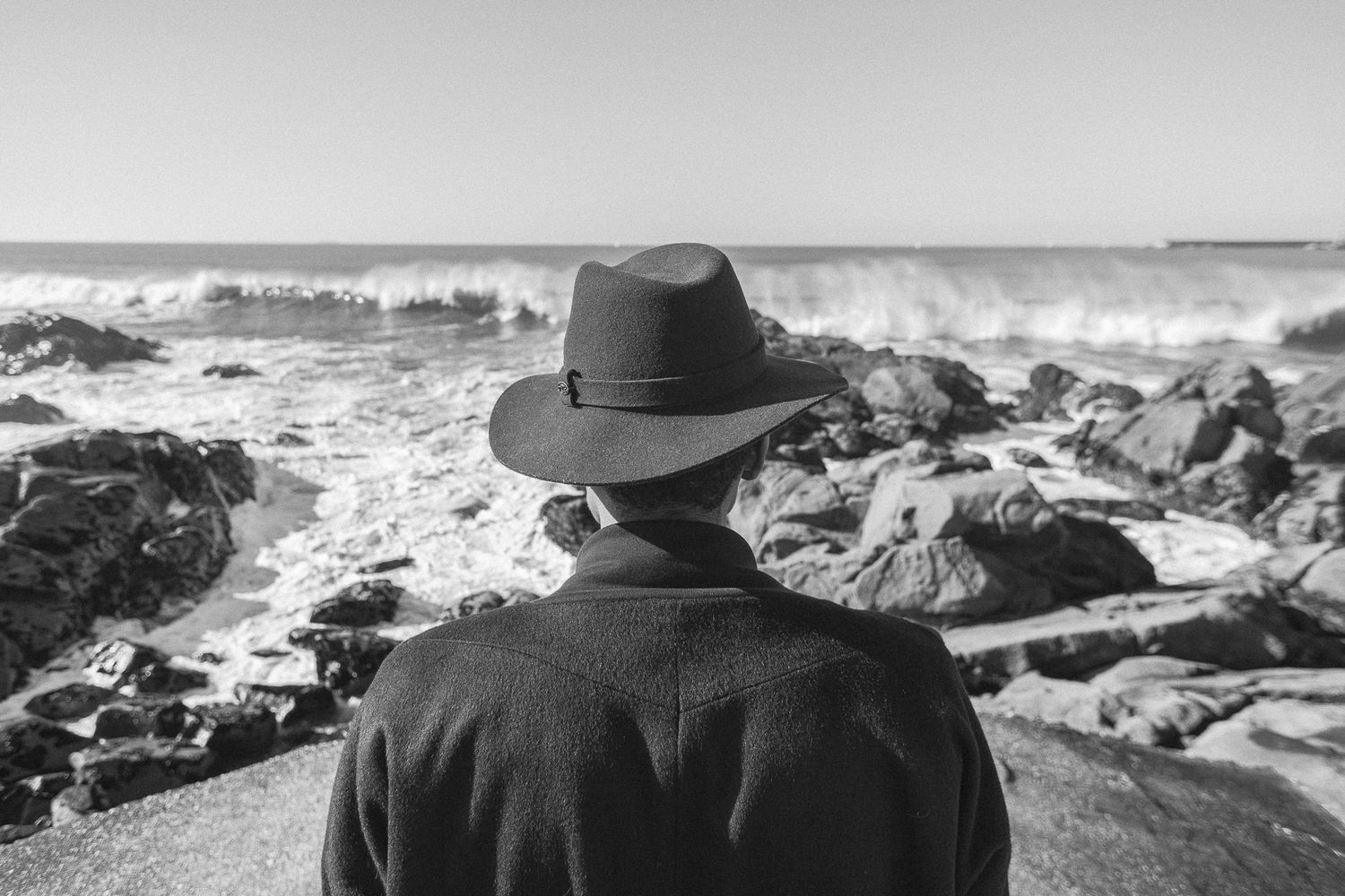 A silhouetted figure in a hat looks out at the crashing ocean waves and rocky coastline on a misty day.
