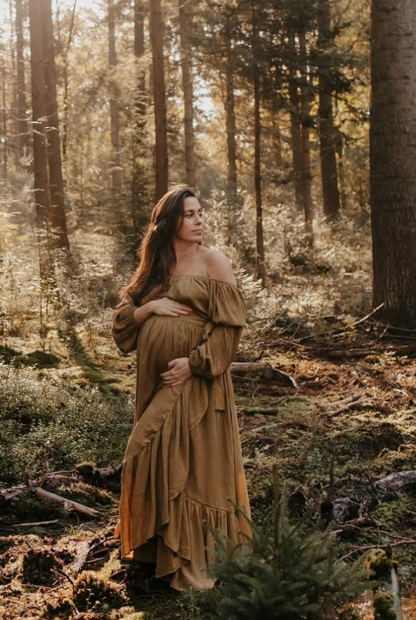 A pregnant woman in a flowing brown dress poses ethereally in a sunlit autumn forest setting.