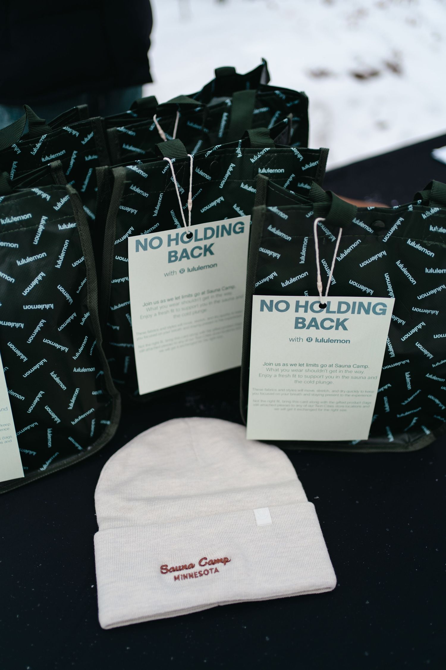 Black patterned gift bags with 'No Holding Back' tags displayed alongside a white beanie hat with red text embroidery.