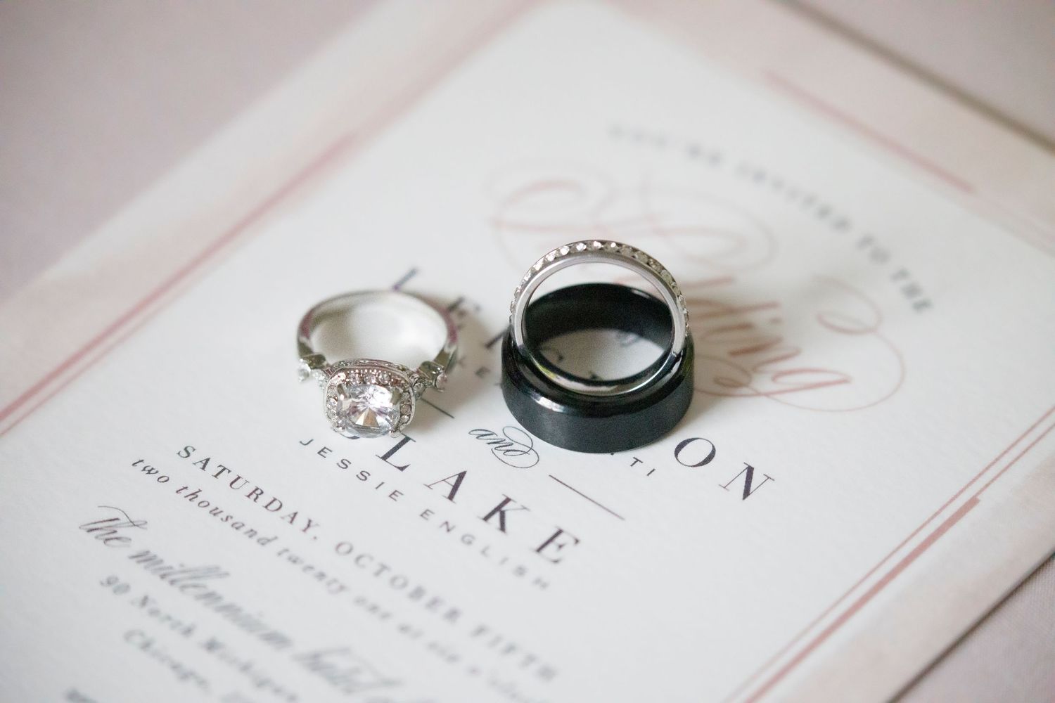 Wedding bands and engagement ring displayed on an elegant wedding invitation in soft, muted lighting.