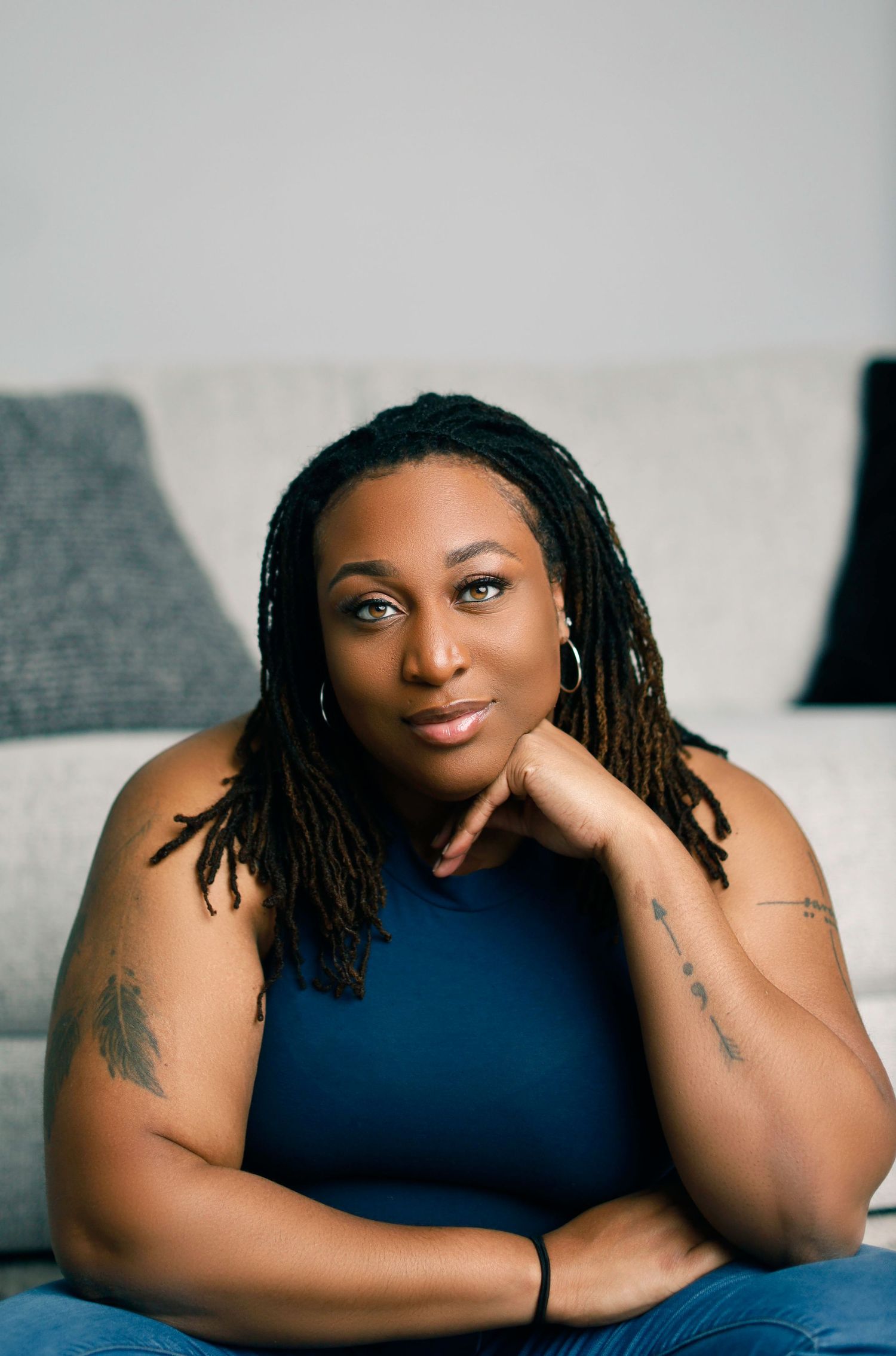 A woman in a navy sleeveless top relaxes on a grey couch with decorative pillows in the background.