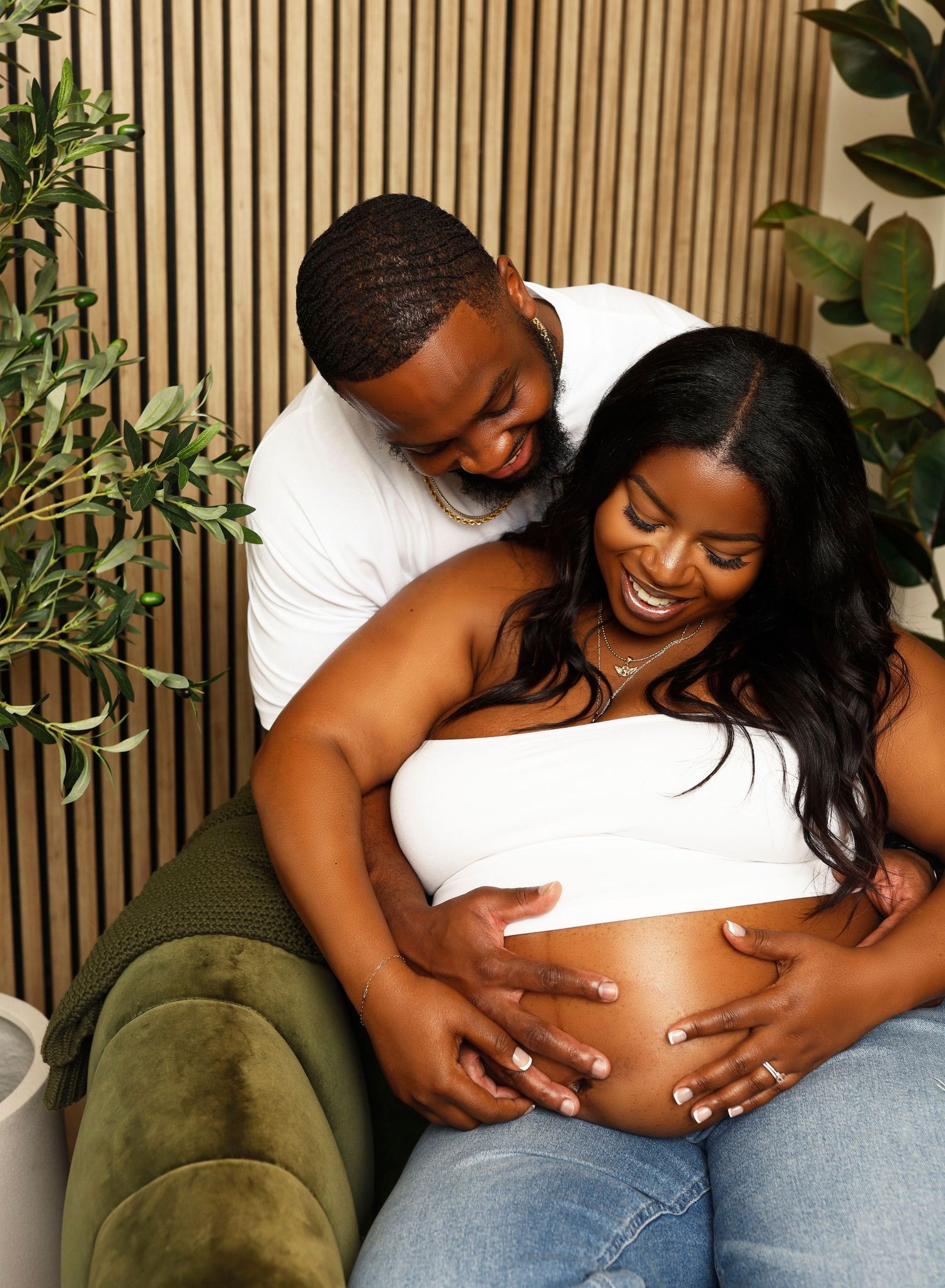 Maternity photo with couple sitting against wooden slat wall, sharing tender moment.