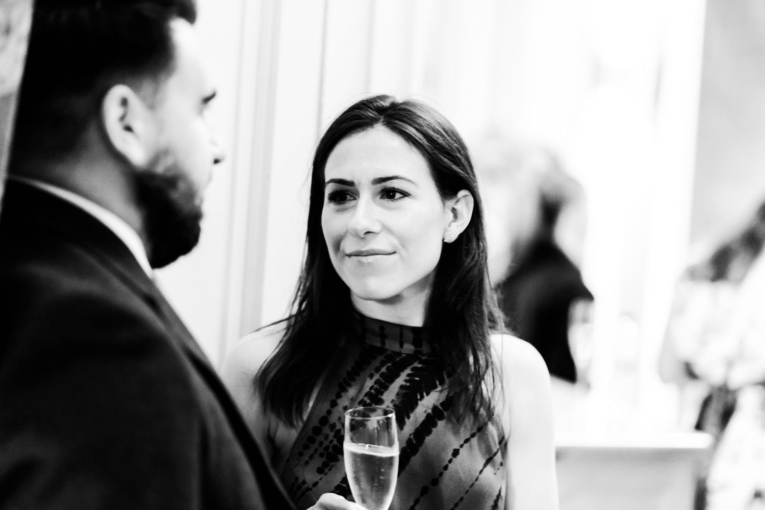A person with dark hair smiles warmly while holding a champagne glass during an elegant black and white conversation.