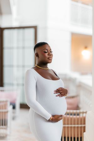 Expectant mother in fitted white dress poses elegantly in bright interior.