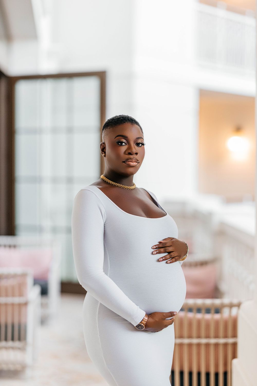 Elegant maternity portrait in white long-sleeved dress against bright interior.