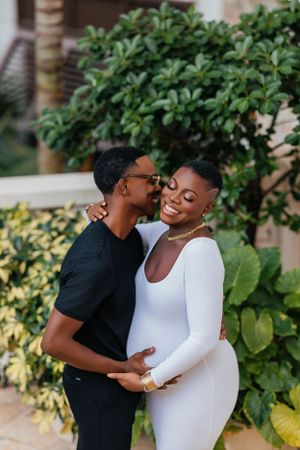 A tender maternity photo shows an intimate moment between a couple against lush greenery.