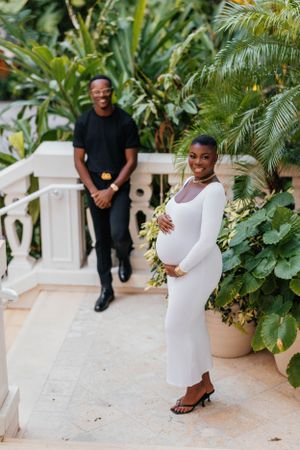 A couple poses for a maternity photo with palm trees and white railings in the background.