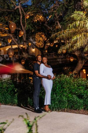 A romantic evening maternity photo with glowing lanterns hanging in trees above the couple.