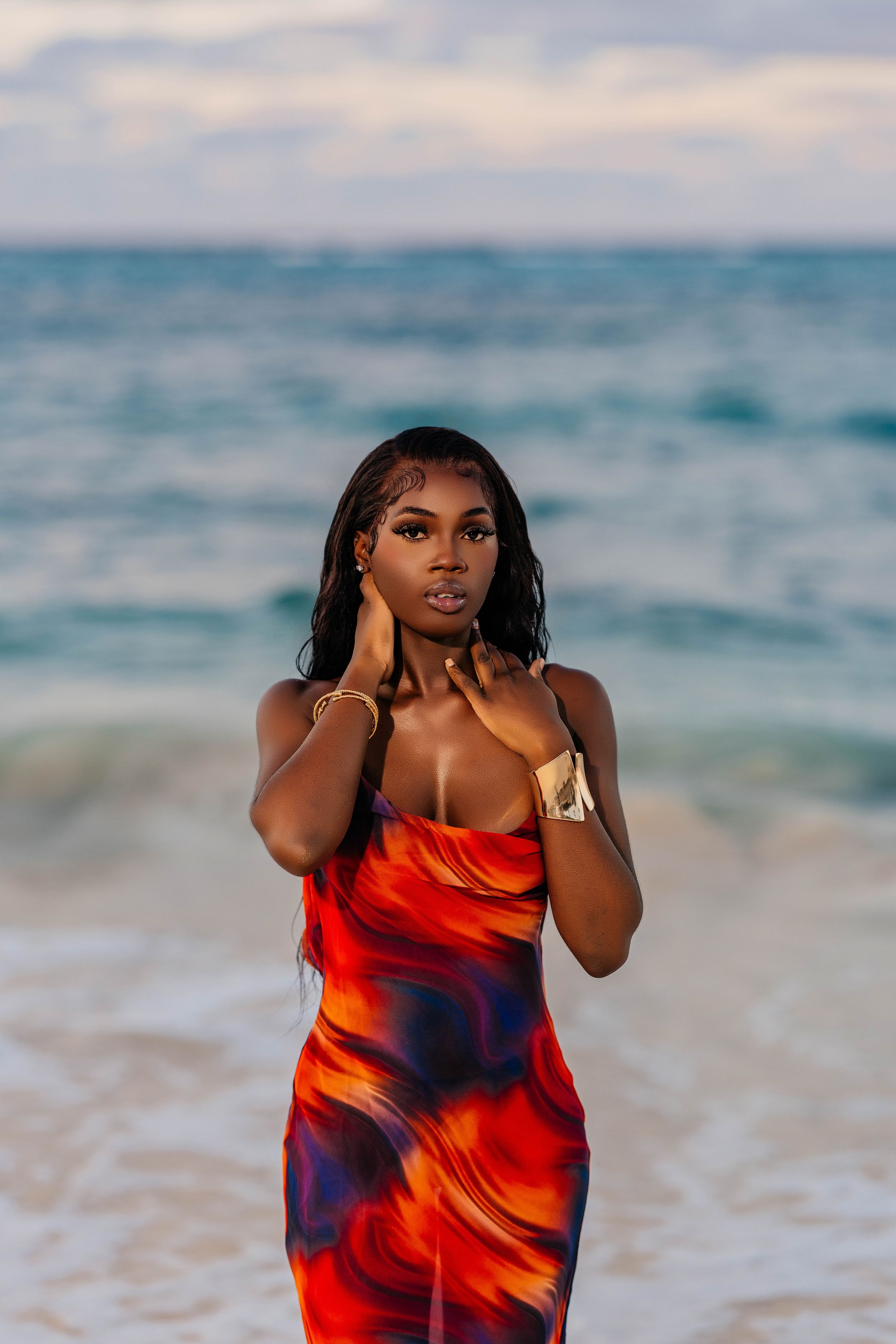 Fashion model in red swirled dress poses on beach at golden hour.