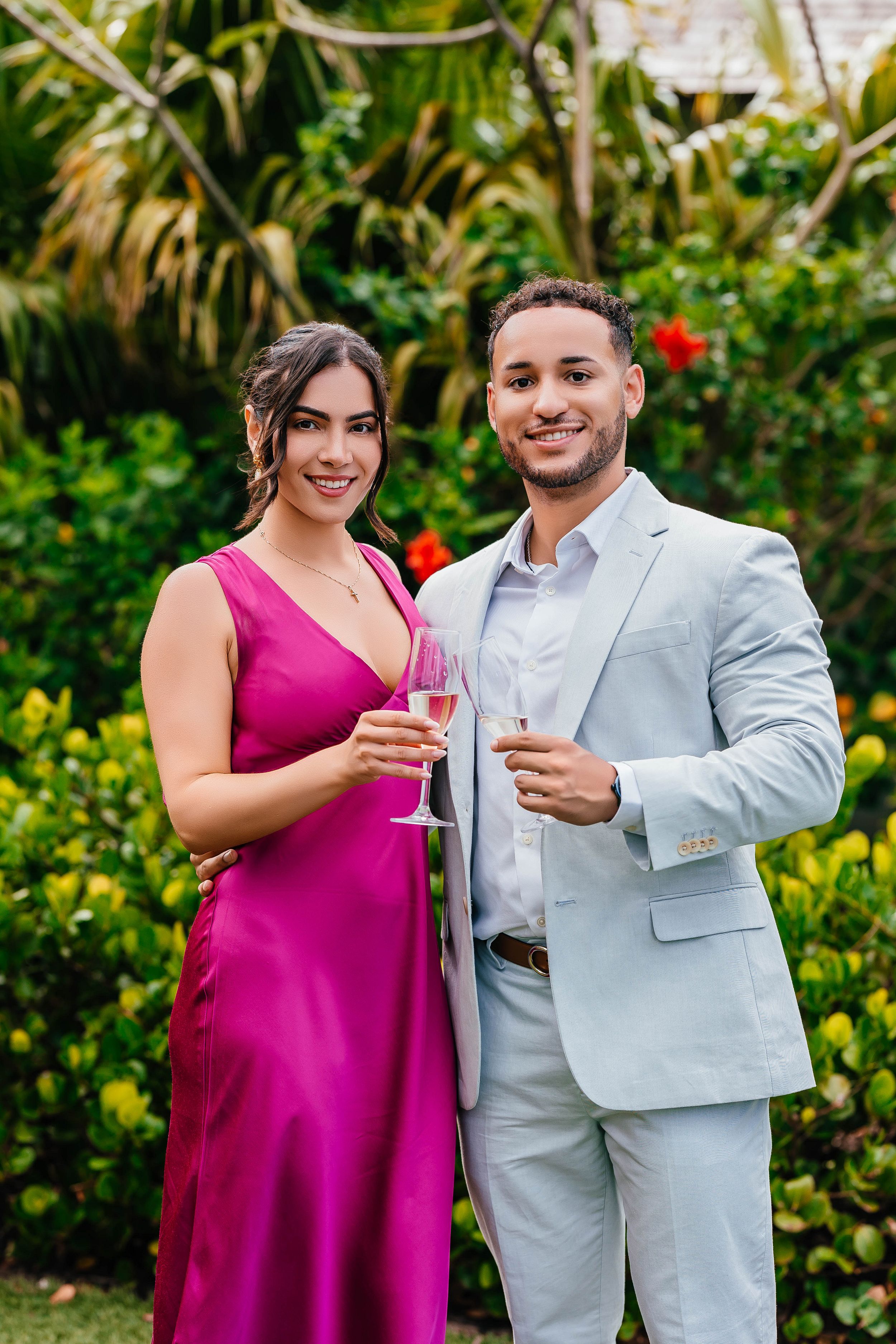 A stylish couple in formal attire toasting with champagne in a tropical garden.