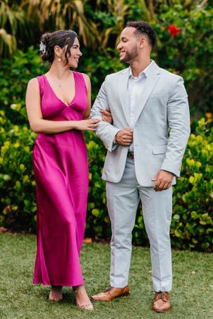 A couple in formal wear smiling at each other against a tropical garden backdrop.