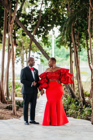 A couple in formalwear poses on a garden path, with the woman wearing a stunning red ruffled mermaid gown.