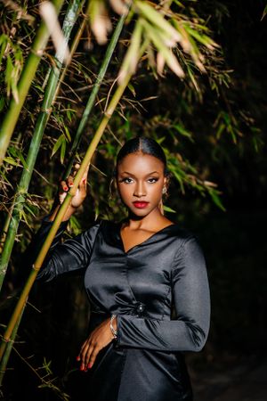 Model in black satin dress poses elegantly among bamboo stalks.