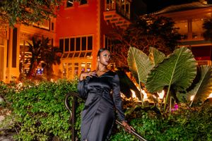Model in sleek black outfit poses against tropical garden and orange building.