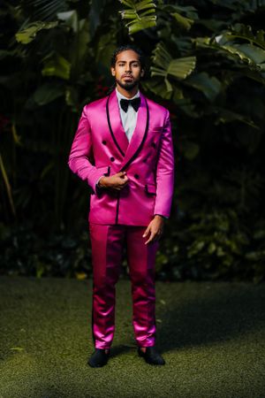 Stylish figure in hot pink formal tuxedo suit poses confidently against lush garden background.