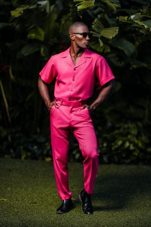 Fashion model in vibrant pink two-piece outfit poses with hands in pockets against tropical foliage.