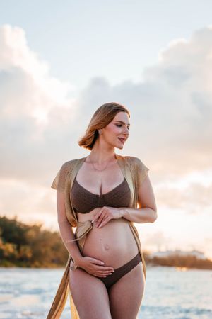 A pregnant woman in a brown swimsuit stands on a beach at sunset.