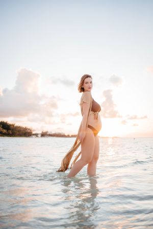 Pregnant woman in brown swimsuit standing in ocean water during golden hour.