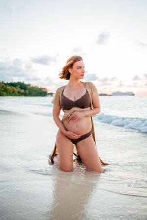 Expectant mother poses in swimwear at sunset on a tropical beach.