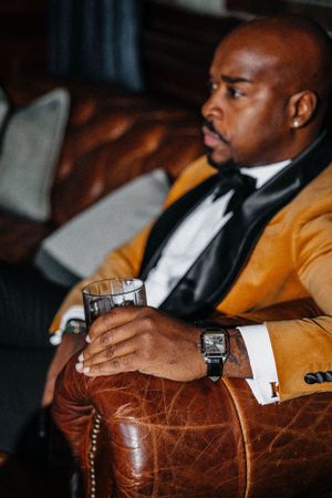 Person in mustard blazer and bowtie sitting on leather chair with glass in hand.