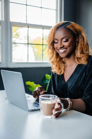 A person with wavy honey-colored hair works on a laptop with coffee at a bright window desk.