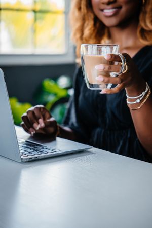 Close-up shot of hands typing on laptop while holding a clear glass mug in a bright office setting.