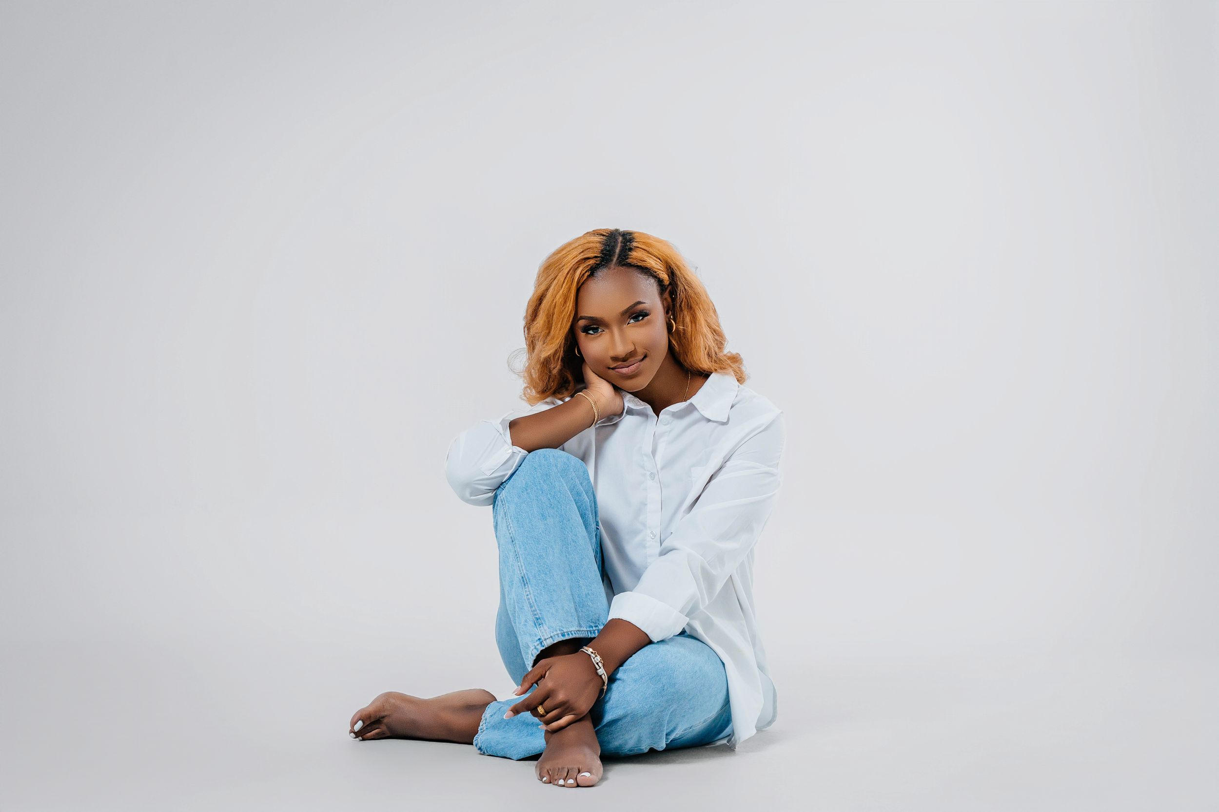 A playful portrait of someone in white shirt and blue jeans sitting cross-legged against white background.