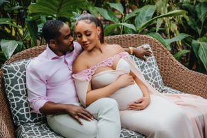 Couple shares intimate moment on wicker furniture during outdoor maternity session.