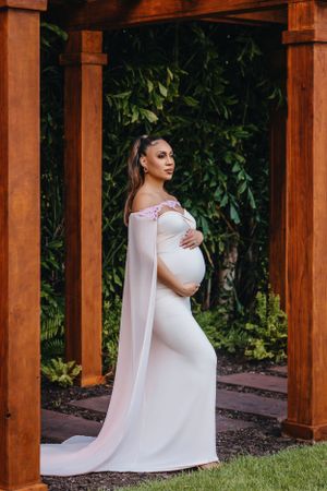 Elegant maternity pose under wooden pergola wearing flowing white gown.